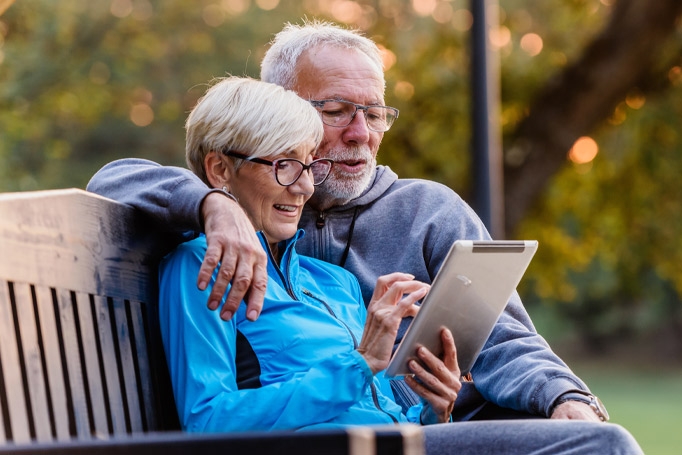 An elder couple wearing jackets sitting on a park bench looking at their tablet