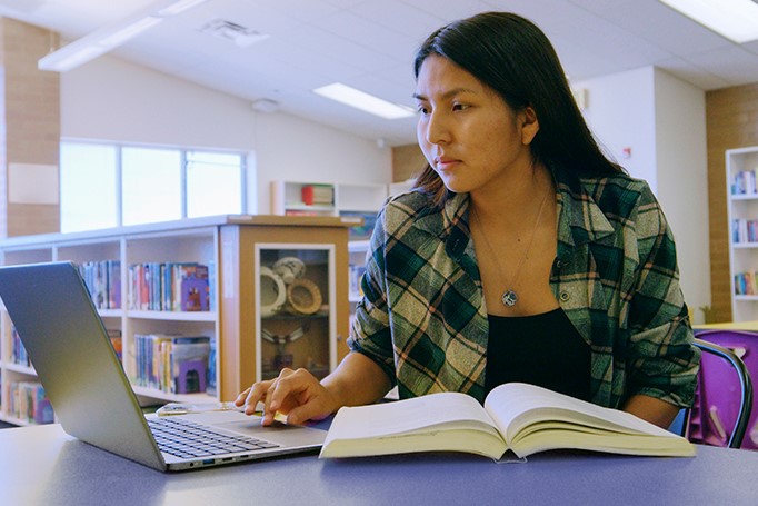 Female student sitting in library with laptop and book researching